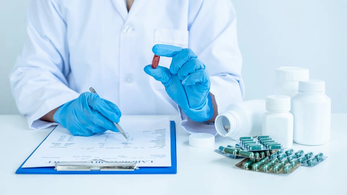 Gloved hands holding a capsule beside a clipboard, medicine bottles and blister packs